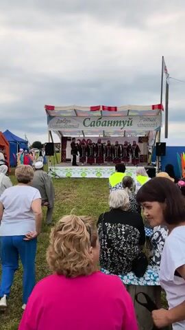 A group of people in traditional clothing are performing on a stage. The stage has a banner that reads "Сабантуй". An audience is watching the performance. The weather appears to be overcast.