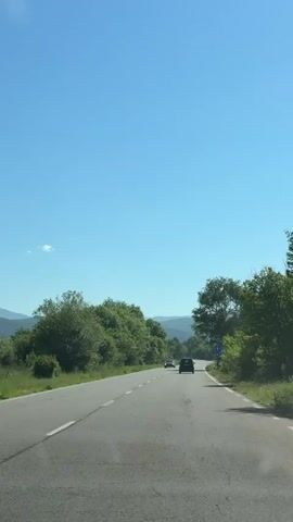 A vehicle drives along a two-lane paved road on a sunny day. Two other cars are seen in the distance, moving in the same direction. The road is bordered by lush green trees and vegetation. A blue road sign is visible on the right side of the road, and a speed limit sign is further ahead. The sky is clear and blue, and distant hills are visible on the horizon.