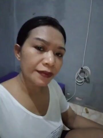 The video shows a close-up of a woman's face as she looks into the camera and speaks. She is wearing a white t-shirt and dark shorts. Her hair is pulled back and she is wearing makeup, including lipstick. The background is out of focus, but appears to be a wall. There is also a purple cushion visible behind her. The lighting is dim.