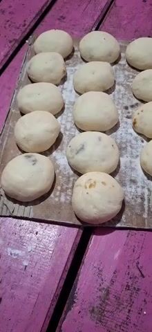 A close-up overhead shot shows a baking sheet lined with parchment paper. Arranged on the parchment paper are approximately 15 small, round, light-colored dough balls. Some of the dough balls have visible darker flecks, suggesting inclusions like raisins or chocolate chips. The baking sheet is placed on a pink wooden surface, which appears to be a table or countertop with visible planks and grain. The lighting is dim, casting shadows and highlighting the texture of the dough.