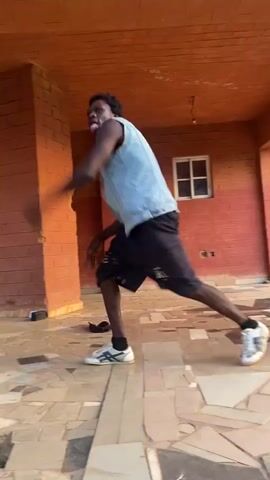 A young man is dancing in an open-air courtyard under an orange brick structure. He is wearing black shorts, a white tank top, and a light blue jean vest. His hair is short and dark. He also wears white sneakers and a white choker necklace.  The dance involves acrobatic movements, including leg extensions while supported by his hands. The floor is tiled with geometric patterns. There is a window visible in the background. A pair of black flip-flops lies on the ground.