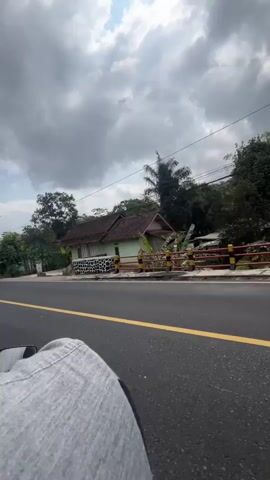 The video shows a view from the perspective of someone riding on the back of a motorcycle. The road is asphalt with a yellow line. On the right side of the road are several houses, some with green walls and tiled roofs. Trees and vegetation are visible behind the houses. A white and green van is driving in the opposite direction on the road. The sky is cloudy.