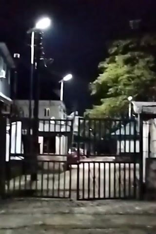 The video shows a night scene in front of a building enclosed by a metal gate. The camera initially focuses on the gate before panning upwards to show the building behind it. The building is a two-story structure, painted a dark color. Two streetlights illuminate the area, casting light on the building, the gate, and a tree on the right side of the frame. There are no people visible in the frame.
