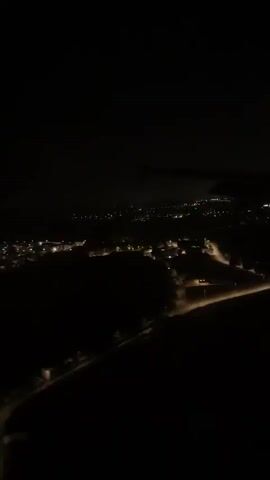 This is a first-person view from an airplane window at night. The camera is looking down at a dark landscape dotted with distant city lights. The wing of the airplane is visible in the upper left corner of the frame, casting a shadow on the scene below. The lights are scattered and appear to be concentrated in small towns or villages. The overall impression is one of a vast, dark expanse punctuated by pockets of human habitation. The video is shaky, suggesting movement, likely the plane in flight.