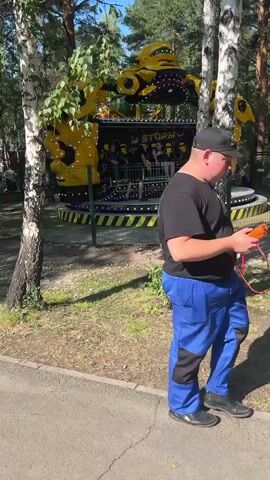 A yellow and black amusement park ride labeled "STORM" operates in a wooded area. The ride has a central platform with rows of seats, on which several people are seated. The ride structure itself is large and yellow, with prominent, angular features resembling mechanical parts. The ride is surrounded by a fence, and trees with white bark are visible in the foreground, casting shadows on the grassy ground. The sky is visible through the trees, indicating it is daytime.