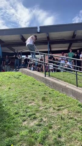 The video begins with a full shot of a young person on a scooter, mid-air, performing a trick over a railing. They are wearing a white t-shirt with a graphic and dark pants. A crowd of spectators, mostly young males, watch from the side, some standing and others seated on benches or a picnic table. The environment appears to be outdoors with a grassy slope in the foreground and trees and a building in the background, suggesting a park or school setting under a bright sky.

The perspective shifts to a medium shot of the scooter rider now on the ground, completing a turn. The rider is still in motion, their body coiled as they prepare for the next action. The crowd continues to watch, with many holding phones to record.

The camera then focuses on the crowd from behind, with the back of someone holding a scooter partially visible in the foreground. The spectators are closely observing the scooter rider, who is out of frame.

Finally, the video shows a different individual, a man with tattoos on his arm, holding a scooter and addressing the crowd. He is wearing a black t-shirt and dark pants. The crowd remains gathered, watching him. The scene is outdoors, with trees and a pathway.