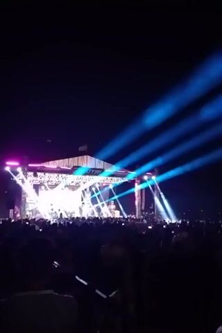 The video shows a concert at night. A stage is illuminated by bright white and blue lights, with a crowd of people in front of it. The crowd is dark, but some individuals are visible in the foreground. The stage has a large screen or banner at the top, with the letters "UTTR" visible. The blue lights are projected upwards into the dark sky, creating a dramatic effect. The scene suggests a lively and energetic event.