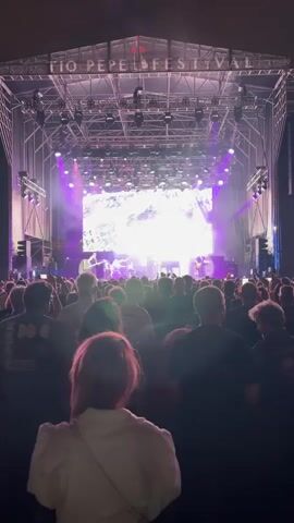 This is a vertical, medium shot of a concert crowd viewed from behind, facing a brightly lit stage. The stage is adorned with a large screen displaying a white and grey abstract pattern, illuminated by bright white and purple lights. Silhouettes of band members are visible on stage, performing under the lights. Above the stage, a banner reads "TIO PEPE FESTIVAL". The crowd, packed closely together, is mostly in silhouette with some individuals partially visible, their faces turned towards the stage. Several people are holding up phones, presumably to record or take pictures of the performance. The atmosphere appears to be that of an outdoor music festival at night.