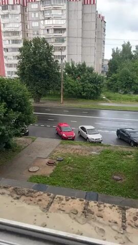 A vertical video captures a cloudy day scene from a window, looking out over a street. In the foreground, the top edge of a window frame and brickwork is visible. Beyond this, a paved walkway leads to a grassy area with manhole covers. Across the street, a multi-lane road has a few cars parked and driving. The road appears wet, suggesting recent rain. Behind the road, a large, multi-story apartment building dominates the background. The building has a light-colored facade with red and white decorative elements along the top. Trees and foliage surround the building and line the street. The sky is overcast and grey.