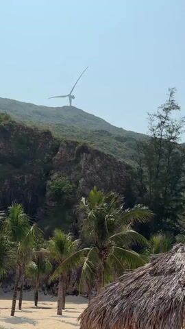 The video shows a sandy beach with thatched-roof umbrellas and lounge chairs, set against a backdrop of green, hilly terrain. Two wind turbines are visible on the hilltops. There are also Asian-style pavilions nestled amongst the vegetation on the hills. The sky is clear and blue.