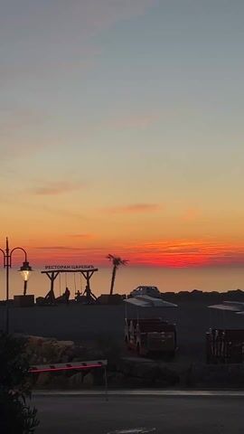 The video shows a picturesque sunset scene. In the foreground, there is a barrier, likely a parking barrier, and two small, red tourist vehicles with white canopies. Behind them, a silhouette of a swing set with two people swinging can be seen, and a sign reads "Ресторан Царевич" (Restaurant Tsarevich) in Serbian. A lone palm tree stands next to the swing set. In the background, the sky is a gradient of colors, transitioning from a pale orange near the horizon to a light pink and blue higher up. The horizon line is a vibrant red, suggesting the sun has just set. A car is parked on the hill behind the swing set. The overall scene is calm and peaceful.