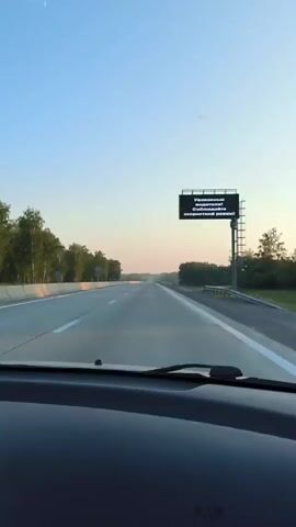 A car is driving on an empty highway during the day. The sky is clear and blue. There are trees on both sides of the road. There is a billboard on the right side of the road with text that reads, "Dear drivers! Please observe the speed limit!" The car is moving forward at a steady pace.