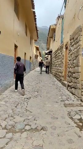 The video shows a narrow, stone-paved street in a town. On either side are buildings of varying heights, mostly with yellow walls and some with visible stonework. In the distance, mountains are visible. A person wearing a black tracksuit and white shoes is walking towards the camera, away from the other two people.  Two other people, a man and a woman, are walking away from the camera down the street toward the mountains. The sky is overcast, providing diffused lighting.