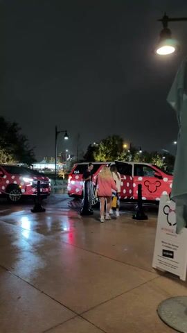 At night on a wet, paved surface, two red Chevrolet SUVs with white polka dots are parked. A man stands near the open rear door of one SUV while a blonde woman enters it. A sign for Minnie Van service is visible in the foreground. A Disney bus is parked nearby. The overall scene appears to be at a Disney theme park or resort. Streetlights illuminate the area.
