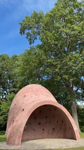 A video shows an outdoor sculpture made of pink bricks, resembling a domed structure with an arched opening. The sculpture is surrounded by grass and a large tree with green foliage. The sky is blue with some clouds. The camera pans slowly around the sculpture, offering a 360-degree view. There are no people or other objects visible in the scene.