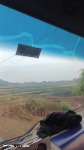 The video shows a view from inside a vehicle, likely a car, moving along a road. The windshield has a blue tint, and there are some cracks visible. A black cloth rests on the dashboard. The outside scene consists of open fields, with a mix of green and brown vegetation. There is a hazy sky above, and distant trees are visible on the horizon. A sticker is attached to the windshield. The video appears to be taken from a low angle within the vehicle.