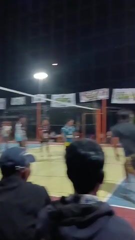 The video shows a volleyball game being played at night. The court is lit by artificial lights, and there are spectators in the foreground, seen from behind. The players are visible on the court, wearing athletic uniforms. The game appears to be in progress, with players serving, setting, and hitting the ball over the net. There are banners visible in the background, suggesting the event may be part of a tournament or league. The atmosphere seems energetic, with the players actively engaged in the match.