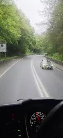 This video shows a first-person perspective of driving a vehicle on a wet, winding road through a forest. The road has double white lines in the center. There are speed limit signs visible, including one for 50 mph. Several other vehicles, including cars and trucks, are on the road ahead. The trees on either side of the road are lush and green, indicating it is likely spring or summer. The weather appears to be overcast and rainy, as evidenced by the water droplets on the windshield and the wet road surface.