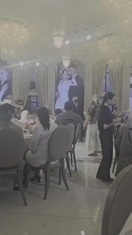 The video captures a wedding reception indoors, likely in Karakol City, Kyrgyzstan. The scene is grand, with large chandeliers and ornate ceiling details. A prominent feature is a large screen displaying a wedding photo of a couple. Guests are seated at round tables, suggesting a formal banquet. Some individuals are standing and interacting, while others are engaged in conversation. The lighting appears to be artificial, indicating an evening or indoor event. The atmosphere seems celebratory, with a sense of gathering and festivity. The attire of the guests suggests a mix of traditional and modern clothing, with some women wearing headscarves. There is no visible text in the video.