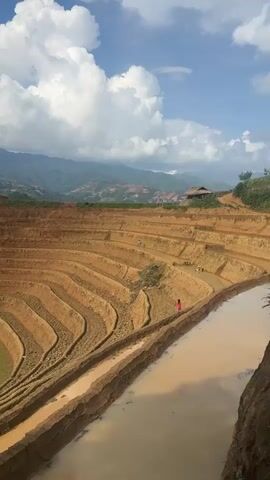 The video presents a panoramic view of expansive terraced fields in Lao Chai, Vietnam, under a bright, partly cloudy sky. The terraces, carved into a steep hillside, display a striking pattern of curved steps, predominantly showing reddish-brown earth, suggesting they are fallow or newly prepared. Some lower sections feature patches of green vegetation, and several terraces are filled with muddy water, reflecting the sky. In the mid-ground, two small figures, likely people, can be seen within the fields, adding a sense of scale. A small, rustic hut is visible on a higher slope in the distance, while a range of mountains forms the backdrop. The scene captures the impressive agricultural landscape and human interaction with the natural environment.