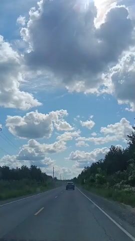 The video shows a view from inside a car driving on a road. The road is paved and has lane markings. Trees and bushes line both sides of the road. The sky is blue with white and gray clouds, and the sun is visible. A dark-colored SUV is driving ahead of the car. The time stamp indicates the video is approximately 5 seconds long.