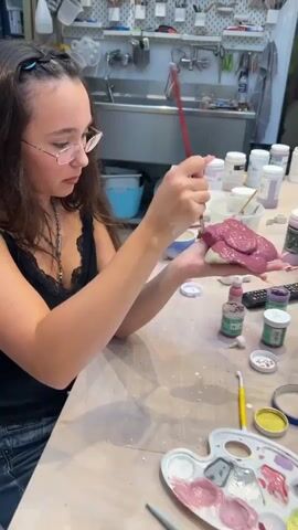 A young woman with glasses and dark hair is sitting at a wooden table, painting a small ceramic object. She is wearing a black top and denim shorts. On the table are various art supplies, including paints, brushes, and other ceramic pieces. In the background, a kiln and shelves filled with art supplies are visible, suggesting a pottery studio. The video then shifts to show other ceramic pieces, including a spherical object with a honeycomb pattern, and various bowls and cups, some with small decorative elements.