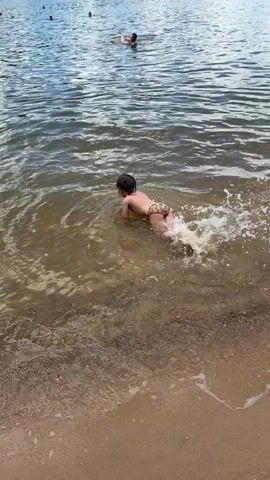 A child in patterned swimming trunks is on their stomach in shallow water at a beach, kicking their legs and moving forward. The water is cloudy and the sandy bottom is visible. In the background, there are other people in the water, a tree line, and a blue sky with clouds.