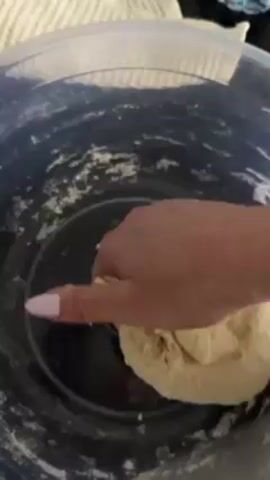 A single hand with pale pink painted nails, adorned with bracelets, kneads dough in a blue plastic bowl. The dough is an off-white color and appears slightly sticky. The bowl, with its translucent lid partially visible, sits on a black countertop next to a folded white and beige striped towel and a partially visible plastic water bottle. Traces of flour are visible inside the bowl. Background music plays.