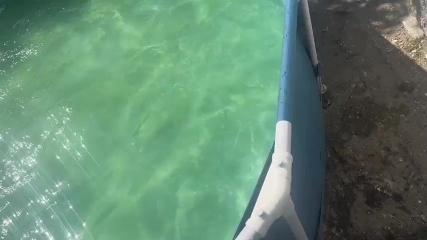 A person is wading into a small above-ground swimming pool. Sunlight glints off the green water. The pool has a blue vinyl liner and a white PVC pipe structure attached to its edge, which appears to be part of a skimmer or filter system. The ground next to the pool is dirt and debris. The person's legs and feet are visible as they enter the water.