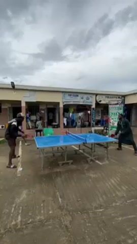 Two men are playing table tennis in an outdoor courtyard on a cloudy day. The ping pong table is blue and silver and sits on a cracked, gray concrete surface. Both men hold paddles and are actively engaged in a rally. The man on the left wears a black jacket, black shorts, and white sneakers. He has a black cap on his head. The man on the right wears a long-sleeved, dark green traditional outfit. Behind them is a row of small businesses. One sign reads "TOP CLEAN" and another displays "NIIN NYSC." People can be seen inside and near the doorways of the shops. Laundry is hanging outside of one of the shops. The overall setting suggests a casual, possibly rural or less affluent location.