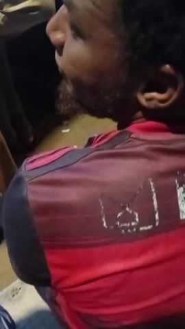 The video shows a close-up of the back of a person wearing a red and black striped shirt. The shirt has some white lettering on it that is partially obscured and illegible. The person's head is turned to the side, and their face is not fully visible. The background is dark and out of focus. The camera movement is slightly shaky.