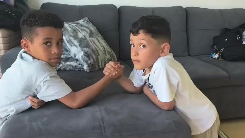 Two young boys are arm wrestling on a grey couch. The boy on the left is wearing a light grey t-shirt and the boy on the right is wearing a white t-shirt. The boy on the left wins the arm wrestling match and smiles at the camera. The boy on the right looks down at his hand. There is a large pillow with a grey and white abstract pattern behind the boys. Luggage is visible in the background on the left side of the image.