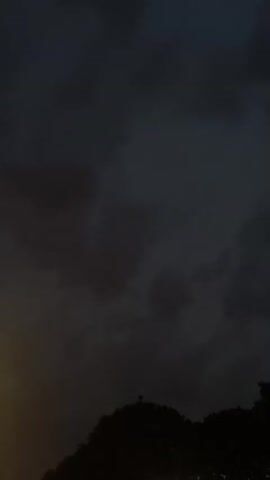 The video shows a time-lapse of dark, stormy clouds moving across the sky, obscuring the sun. Silhouetted against the clouds are the tops of trees and a hill, with occasional faint lights visible at the bottom of the frame, possibly from streetlights or buildings. The overall mood is ominous and foreboding.