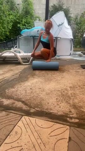 A woman is performing a squat with a foam roller in front of her. She is wearing a black and blue swimsuit. The foam roller is blue and the mat is black. The scene is outdoors in a backyard. There is a swimming pool in the background, along with some lawn furniture and plants. The woman is facing the camera and smiling.