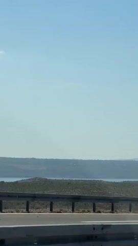 A dark colored sedan drives on a highway next to a body of water. The landscape is hilly and covered in green shrubbery. The sky is clear and blue with a few wispy clouds. A power tower is visible in the distance.