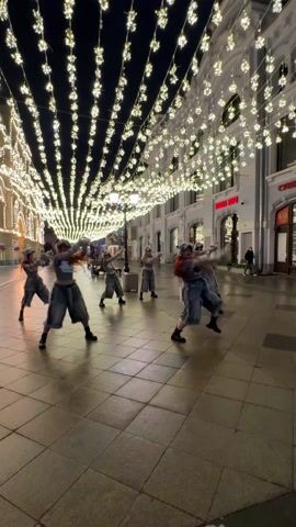 A group of dancers, wearing matching outfits consisting of grey crop tops, baggy pants with faux fur trim, boots, and hats with faux fur and tails, are performing a choreographed routine on a city street at night. The street is paved with light-colored tiles and is lined with tall buildings, some with red signage. Strings of decorative lights hang above the street, creating a bright, festive atmosphere. The dancers move in unison, facing the camera for a portion of the video and then turning to show their backs.  A few pedestrians can be seen in the background.