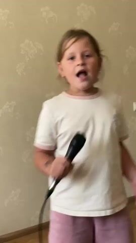 This video shows a young girl, likely of European descent, holding a microphone and singing. She is standing in what appears to be a residential indoor setting, possibly a living room or bedroom, indicated by the floral patterned wallpaper and a wooden floor. The lighting suggests it could be daytime.

The girl is wearing a white t-shirt with pink trim and pink shorts. She has blonde hair and is looking directly at the camera with an expressive face, her mouth open as she sings. She appears to be enjoying herself, with a playful and animated demeanor. Her right hand is holding the microphone close to her mouth, while her left arm is extended slightly to her side.

The background features a light-colored wallpaper with a repeating pattern of stylized flowers in a slightly darker shade. A white electrical outlet is visible on the wall to the right of the girl. The overall impression is of a casual and lighthearted moment, perhaps a karaoke session or practice for a performance. The video captures the girl in various stages of singing, with her mouth open and closed, and with different hand gestures. The audio appears to be of the girl singing, though the exact song is not identifiable. The location context is Gulkevichi, Russia.