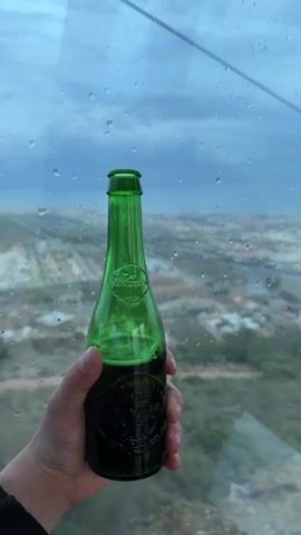 A hand holds a green glass bottle of dark soda against a rain-speckled glass window, revealing a cityscape stretching towards the ocean under a cloudy sky. A cable car passes in the background.