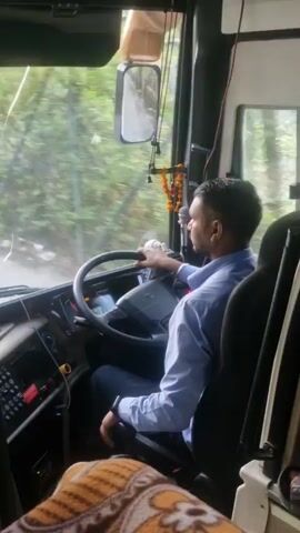 The video shows the interior of a bus, with a driver in the driver's seat. The driver is a man with dark hair and a beard, wearing a light blue collared shirt. He is holding the steering wheel and looking forward. Outside the bus window, trees and a road are visible. The bus appears to be in motion. There is a clock on the dashboard that reads 6:46. There are also some objects in the foreground, including a blanket and a bottle.