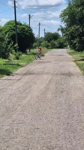 A young child wearing an orange shirt and blue shorts rides a bicycle down a dirt road. The road is lined with trees and grass, and there are power lines running overhead. The sky is blue with some clouds. The child is pedaling and appears to be moving at a moderate speed. The camera is positioned behind the child, following them as they ride. The video is short and appears to capture a simple moment of childhood activity.