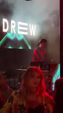 Here's a detailed description of the video:

**Scene:** The video takes place in what appears to be a nightclub or concert venue. The atmosphere is dark, with lighting effects and smoke contributing to the ambiance.

**People:**
*   **DJ:** A person with short, dark hair is positioned behind a DJ setup. He is wearing a red and black striped long-sleeved shirt and headphones. He appears to be operating the mixing equipment and possibly speaking into a microphone.
*   **Audience:** Several people are visible in the foreground, suggesting the presence of an audience. At least one woman with bright red hair is seen near the front.

**Objects:**
*   **DJ Setup:** A DJ console with mixing boards, turntables, or other related equipment is present.
*   **Screen/Backdrop:** Behind the DJ, there is a large screen displaying the text "KC DREW" in white letters with a geometric design in teal and white.
*   **Lighting:** Various lighting effects are in use, including green laser beams and stage lights that cast different colors (e.g., pink).

**Actions & Interactions:**
*   **DJ'ing:** The DJ is actively involved in mixing music, as indicated by his hand movements on the equipment.
*   **Audience Interaction:** The people in the foreground are moving and dancing, implying they are enjoying the music and the atmosphere.

**Environment:**
*   The venue has some floral decorations.
*   Smoke or haze effects are present, enhancing the visual impact of the lighting.

Overall, the video captures a scene from a live music event featuring a DJ performing for an audience in a nightclub-like setting.