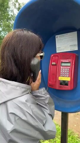 A woman with shoulder-length brown hair is standing in a blue, open-air phone booth. She is wearing a gray hooded jacket and white-framed sunglasses with blue mirrored lenses. She is holding a black telephone receiver to her left ear with her right hand and speaking into it. She is smiling and looking to her right. A red payphone with a digital display and a keypad is mounted on the interior wall of the phone booth to her right. The phone booth is outdoors, with green grass and trees visible in the background. The woman's sunglasses reflect the environment, showing a blurred image of trees and sky. The video cuts between the woman speaking and reacting with a wide smile and closed eyes.