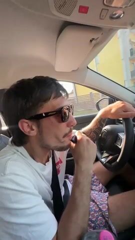 The video opens with a close-up of a man driving a car. He is wearing sunglasses and a white t-shirt with a red graphic. His left hand is on the steering wheel, and his right hand is near his face, holding an object that he appears to be smoking. The car's infotainment screen is visible, displaying "SOS FÜHRER SOS 01:46" with several icons below, including a phone, "Mode," "Keypad," and "Add Emergency Contact." The interior of the car is dimly lit, with sunlight streaming through the windshield. The scene then shifts to a wider shot of the man driving. The car appears to be moving on a street with buildings and trees visible outside the windows. The man briefly turns his head towards the camera and says "Ciao." The video ends with the man continuing to drive, with his hands on the steering wheel and the car in motion.