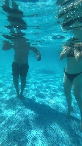The video shows an underwater view of a swimming pool. Two people are visible in the water. On the left, a person wearing dark shorts stands waist-deep. They are holding a white inflatable object in their hands. On the right, a person wearing a bikini is partially visible, lying on their side and holding a similar white inflatable. The pool floor is tiled with small blue and white squares. Sunlight refracts through the water, creating dappled patterns on the pool floor. The footage is shot from a low angle, looking upwards towards the surface.