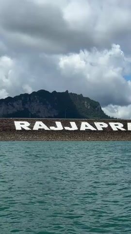 A wide shot shows a body of water with choppy waves in the foreground. In the background, a large dam wall with the white letters RAJJAPRABHA painted across it is visible. Behind the dam, a lush green mountain range with rocky cliffs rises against a cloudy sky. The clouds are varied, with some dark and heavy and others bright and fluffy. There are faint indications of roads and possibly vehicles on top of the dam wall. The overall scene suggests a scenic, natural landscape with a man-made structure.