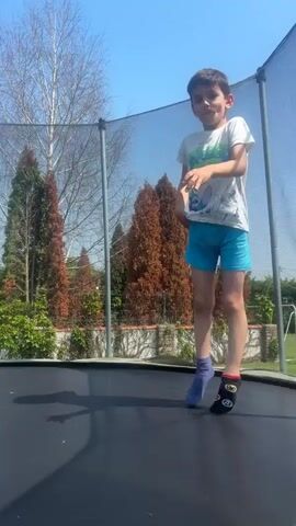 A young boy is actively jumping on a black trampoline outdoors under a clear blue sky. He is wearing a white t-shirt with a blue and green graphic, light blue shorts, and mismatched socks – one purple, the other black with a red heel and a yellow graphic (possibly "27" or an emoji). He appears focused and possibly counting his jumps, saying "1, 2, 3..." at the beginning of the video.

The setting is a sunny backyard or garden, with various trees and shrubs visible in the background. Some trees are bare, indicating either late winter or early spring, while others are evergreen conifers. A safety net surrounds the trampoline. The bright sunlight casts a clear shadow of the boy on the trampoline's surface. The activity takes place in Neofit Rilski, Bulgaria.