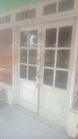 The video shows a close-up, slightly shaky shot of a pair of old, white double doors with glass panes. The doors appear to be made of wood and have a classic design with multiple smaller rectangular panes above solid bottom panels. The door handles are visible, as well as two padlocks securing the doors together. The doors are set within a white frame, and a light green trim is visible along the top. The flooring in front of the doors appears to be tiled. The glass panes reflect the outside environment, though it is difficult to discern specific details due to the lighting and quality of the recording. The overall impression is of an exterior view of a building's entrance.