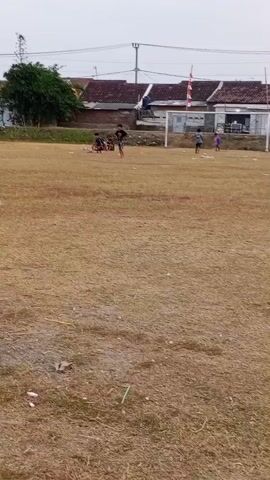 The video shows a blurred, outdoor scene, likely a sports field, with dry, brown grass. Several people are visible, some appearing to be children, scattered across the field. One group is near a soccer goal, and another group is sitting on a bench under a shelter. A bicycle is parked to the right of the goal. In the background, there are buildings with reddish-brown roofs and utility poles. The sky is overcast. The camera appears to be moving, causing the motion blur. The overall impression is of a casual, possibly recreational activity taking place on a somewhat neglected field.