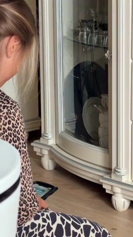 A woman with long brown hair, wearing a leopard-print outfit, is kneeling on a wooden floor in front of a glass-fronted cabinet. She is holding a tablet computer in her hands. The cabinet contains plates and glassware. A white trash can is visible to the left of the frame.
