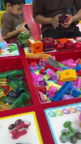 A boy wearing a brown t-shirt is playing with building blocks and a toy tank. An adult, wearing a brown t-shirt and a black bag, is also present and interacting with the blocks. The scene is set in a play area with various colorful building blocks, tubes, and toy vehicle pictures on display.